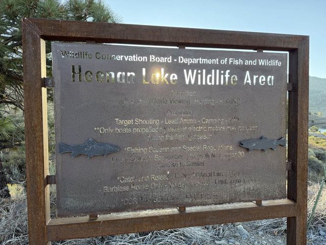 A weathered steel sign with two cut-steel trout: Wildlife Conservation Board, Department of Fish and Wildlife, Heenan Lake Wildlife Area, and below it the permitted uses and the fishing season.