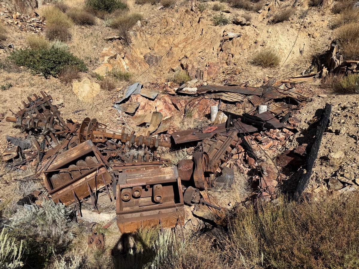 Collapsed machinery on a broken rock slope: a cast iron flywheel, a heavy worm shaft, sheets of corrugated iron and burned timber lying among sagebrush.