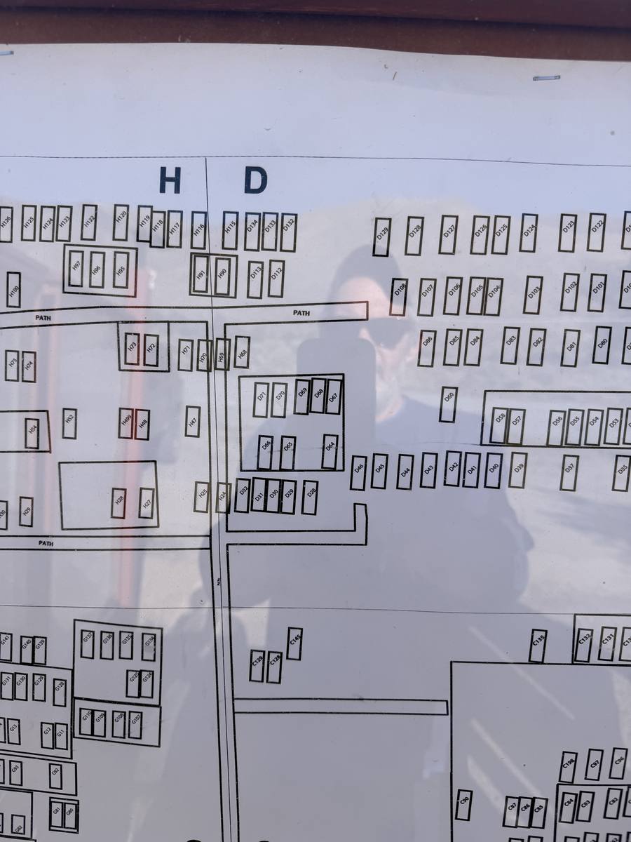 A cemetery plat board under glass: rows of numbered rectangles in lettered sections, with paths between them, and the shadow of the photographer across it.