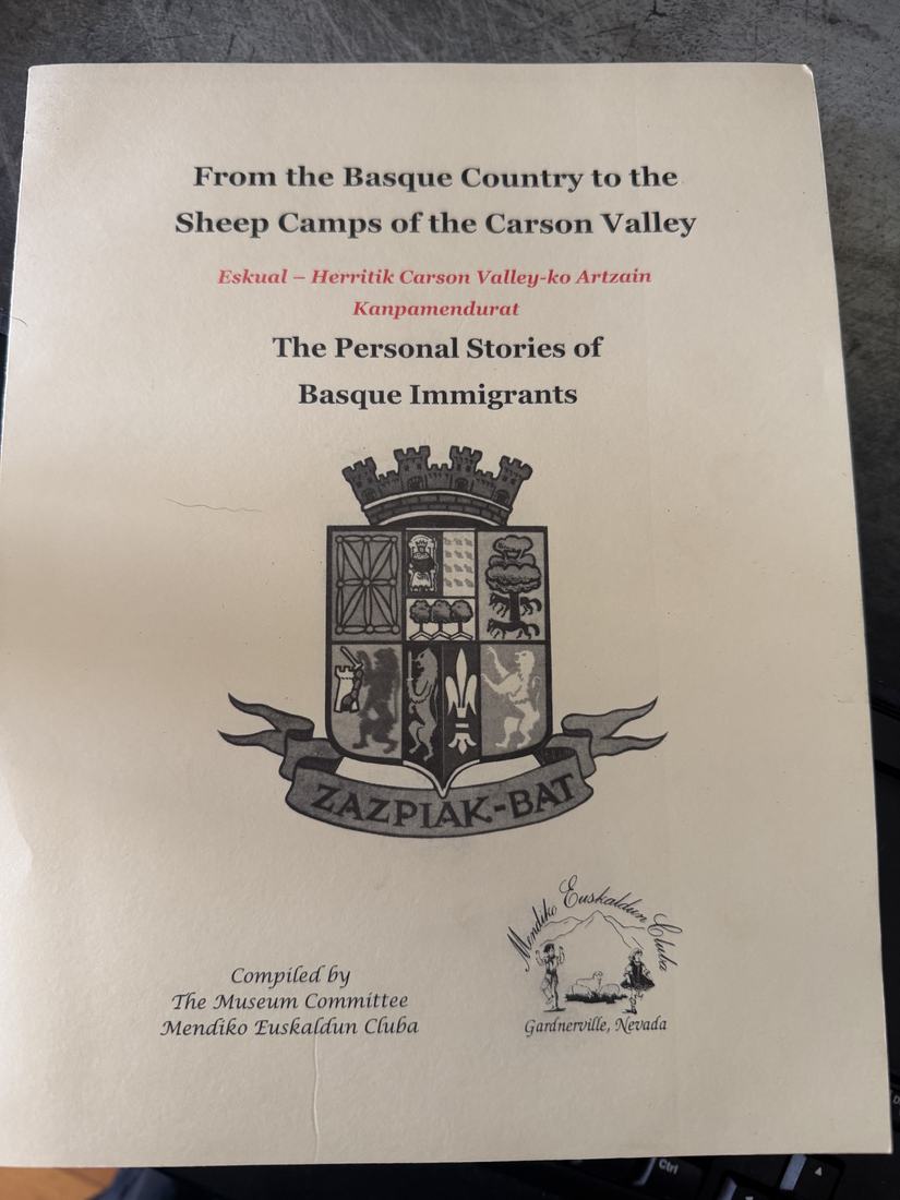 A soft-cover book lying on a table: From the Basque Country to the Sheep Camps of the Carson Valley, The Personal Stories of Basque Immigrants, with the Zazpiak-Bat arms, compiled by the Museum Committee, Mendiko Euskaldun Cluba, Gardnerville, Nevada.