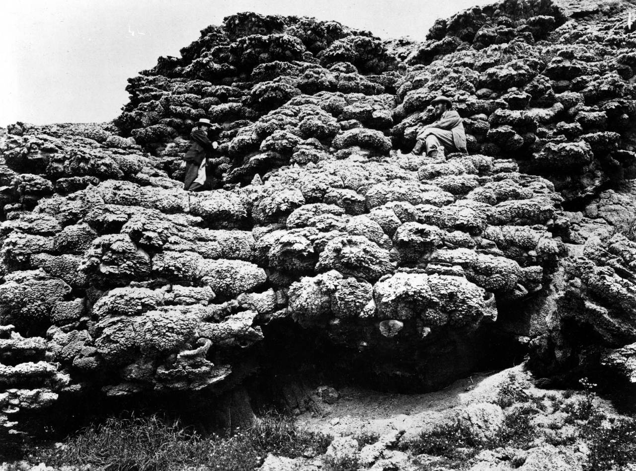 A nineteenth-century photograph of a rough, knobbed mass of pale rock many times a man&rsquo;s height. One man in a hat stands on it at the left; a second sits higher up at the right, wrapped in a coat, looking at the camera.