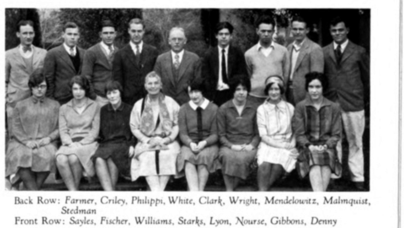 A group photograph of seventeen people in two rows; an older woman in a patterned scarf sits at the centre of the front row.
